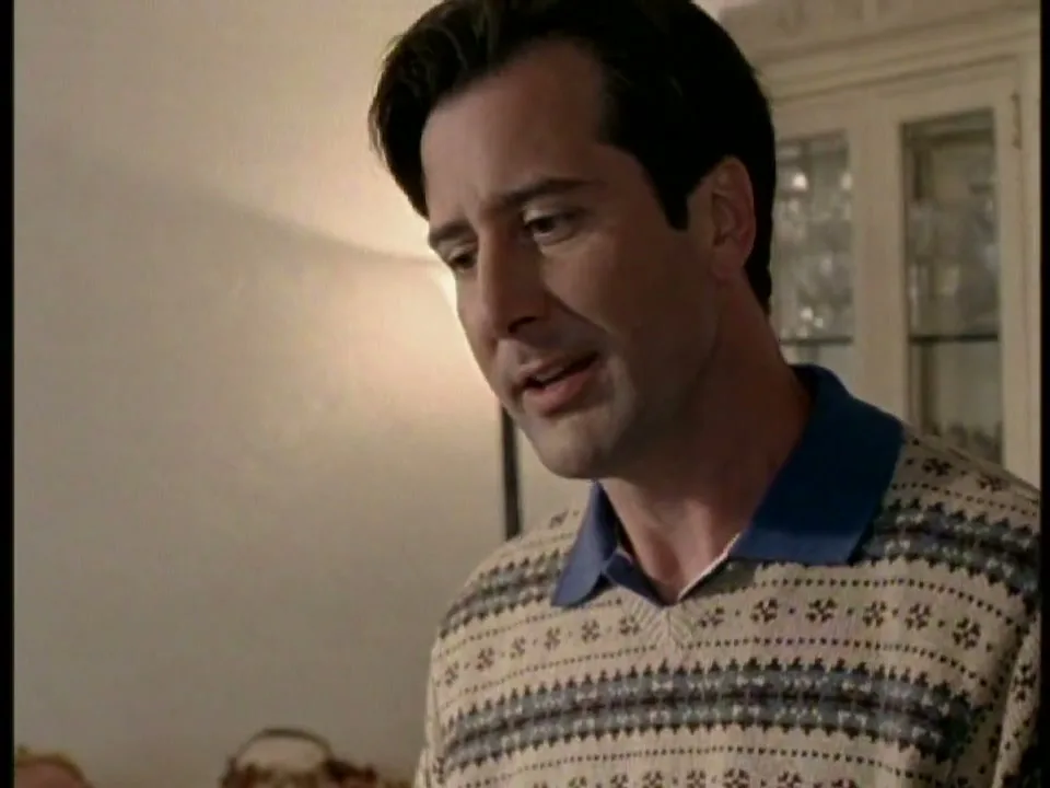 A man in a patterned sweater looks down and smiles, with a woman partially visible in the foreground, in an interior setting.