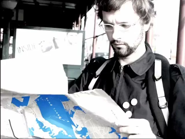 A man with glasses and a beard, wearing a black jacket, looks at a map with blue highlights, appearing confused at a bus stop.