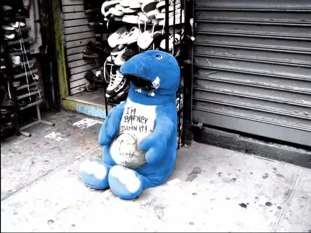A blue stuffed dinosaur toy, with "I'M BARNEY DAMN IT!" written on its belly, sits on a sidewalk in front of a closed store.