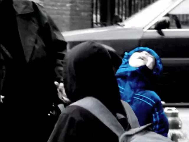 A child in a blue jacket looks up, while an adult and another child, all in black and white, stand nearby on a city street.