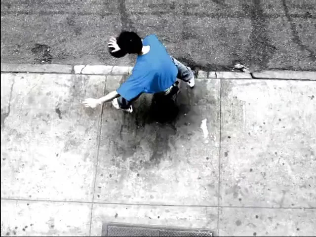 A person in a blue shirt and black hat is seen from above, walking on a sidewalk next to a street, with a color splash effect.