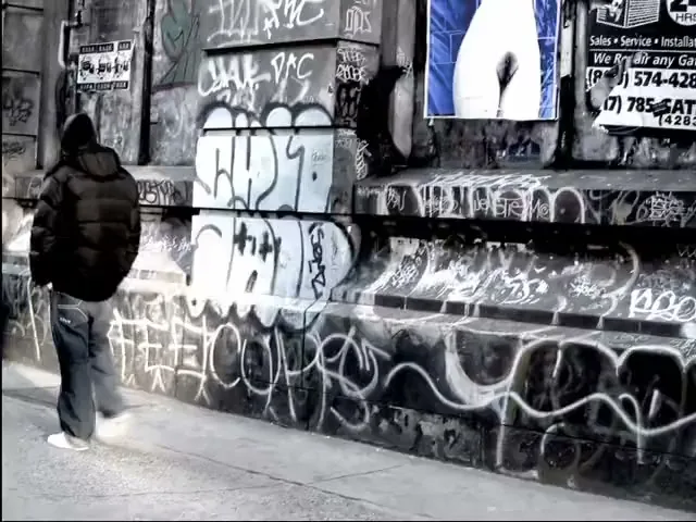 A person in a black puffer jacket and jeans walks past a graffiti-covered wall with posters in a city street.