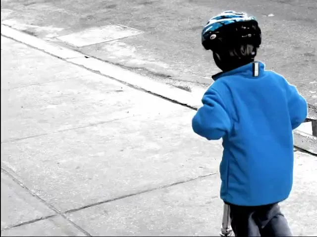 A young boy in a blue jacket and helmet rides a scooter away from the camera on a grey sidewalk.