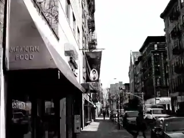 A black and white street scene with a red awning and a black flag with a white symbol, as the camera moves forward.