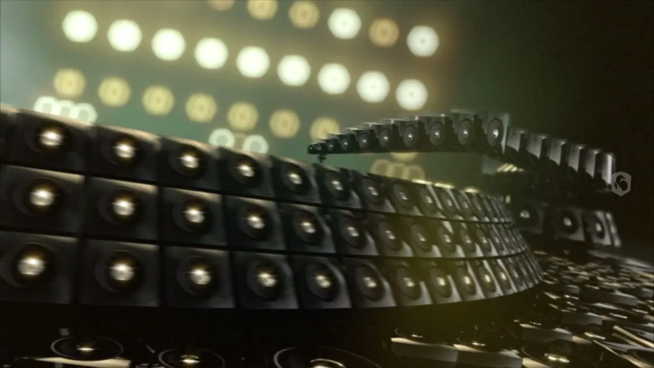A wide shot shows a large, curved wall of speakers with many individual speakers, with a bright, out-of-focus background of lights.