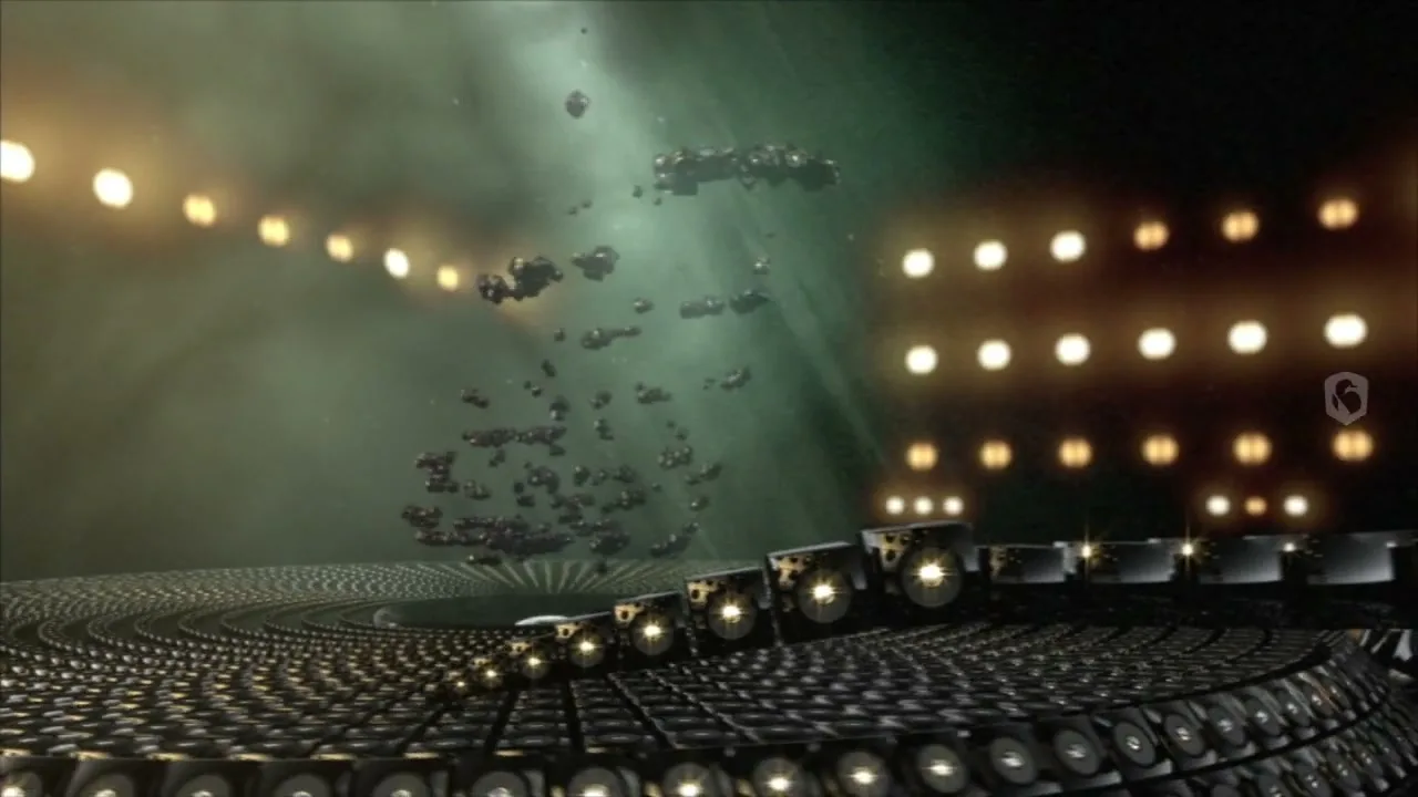 A wide shot of a circular stage made of speakers, with a bright light in the center, as more speakers emerge and swirl around.
