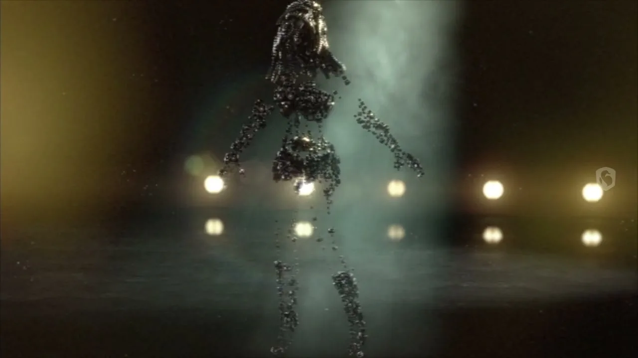 A female figure made of small, dark, metallic spheres forms and dances in a spotlight on a dark stage with blurred lights in the background.