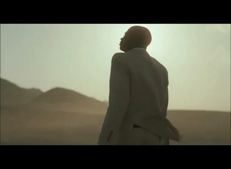 A man in a suit stands in a desert landscape, looking up towards the bright sun, with mountains in the background.