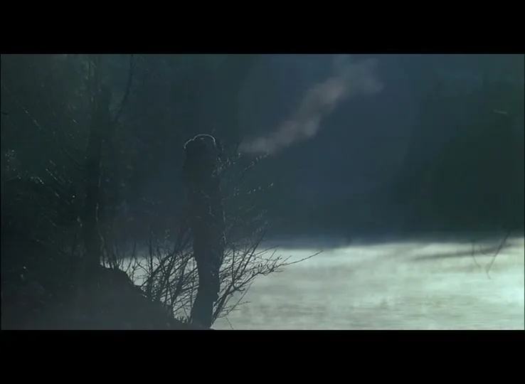 A lone figure stands silhouetted by a misty lake, exhaling visible breath into the cold air at dawn.