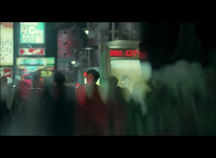 A blurry shot of a woman in a red dress walking through a crowded city street at night, with neon signs in the background.
