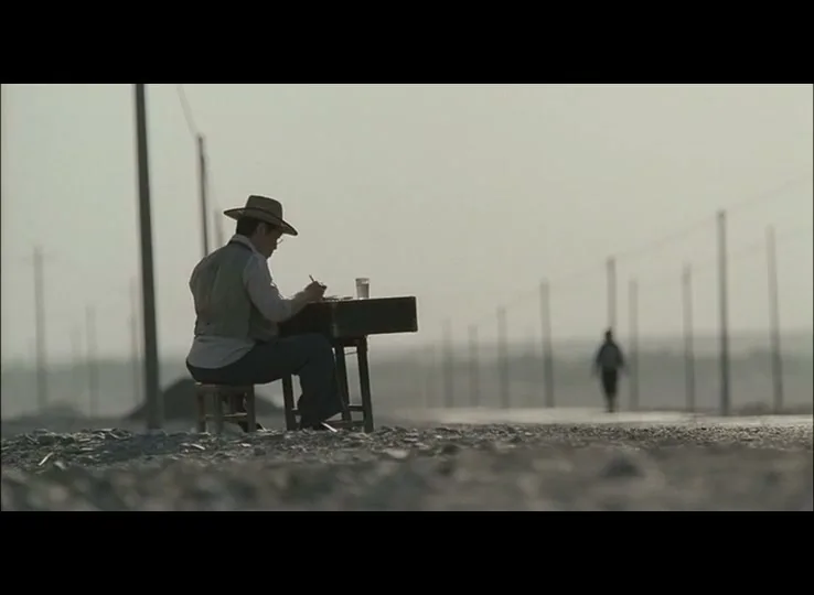 A man in a hat sits at a desk on a dirt road, painting, while another person walks away in the distance under a hazy sky.
