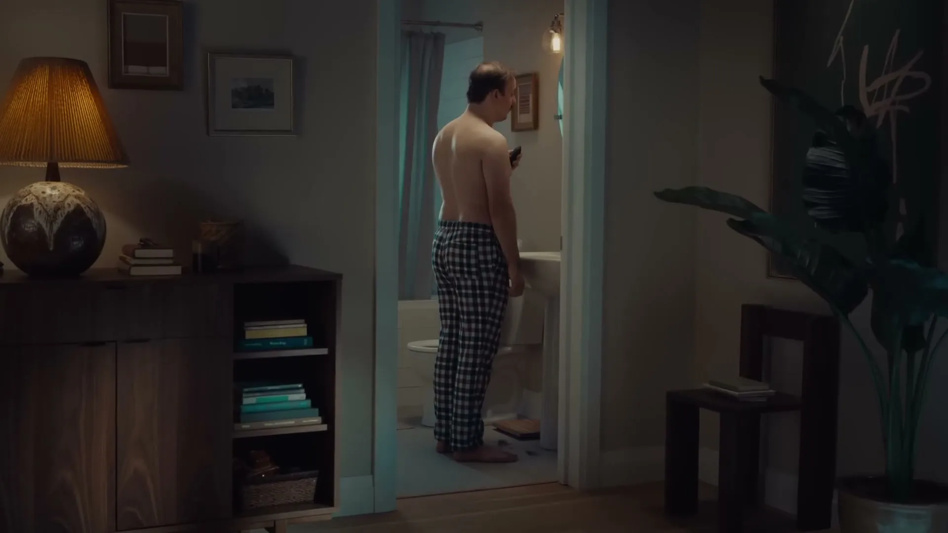 A man in plaid pajama pants stands shirtless in a bathroom doorway, looking down at a trimmer in his hand, in a dimly lit home.