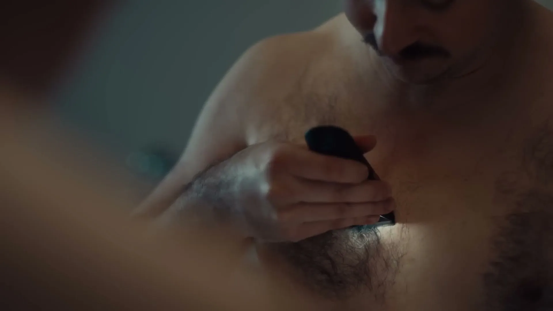 A small, sad-looking hairball with googly eyes sits on a tiled bathroom floor, surrounded by scattered hair, as a man shaves his chest.