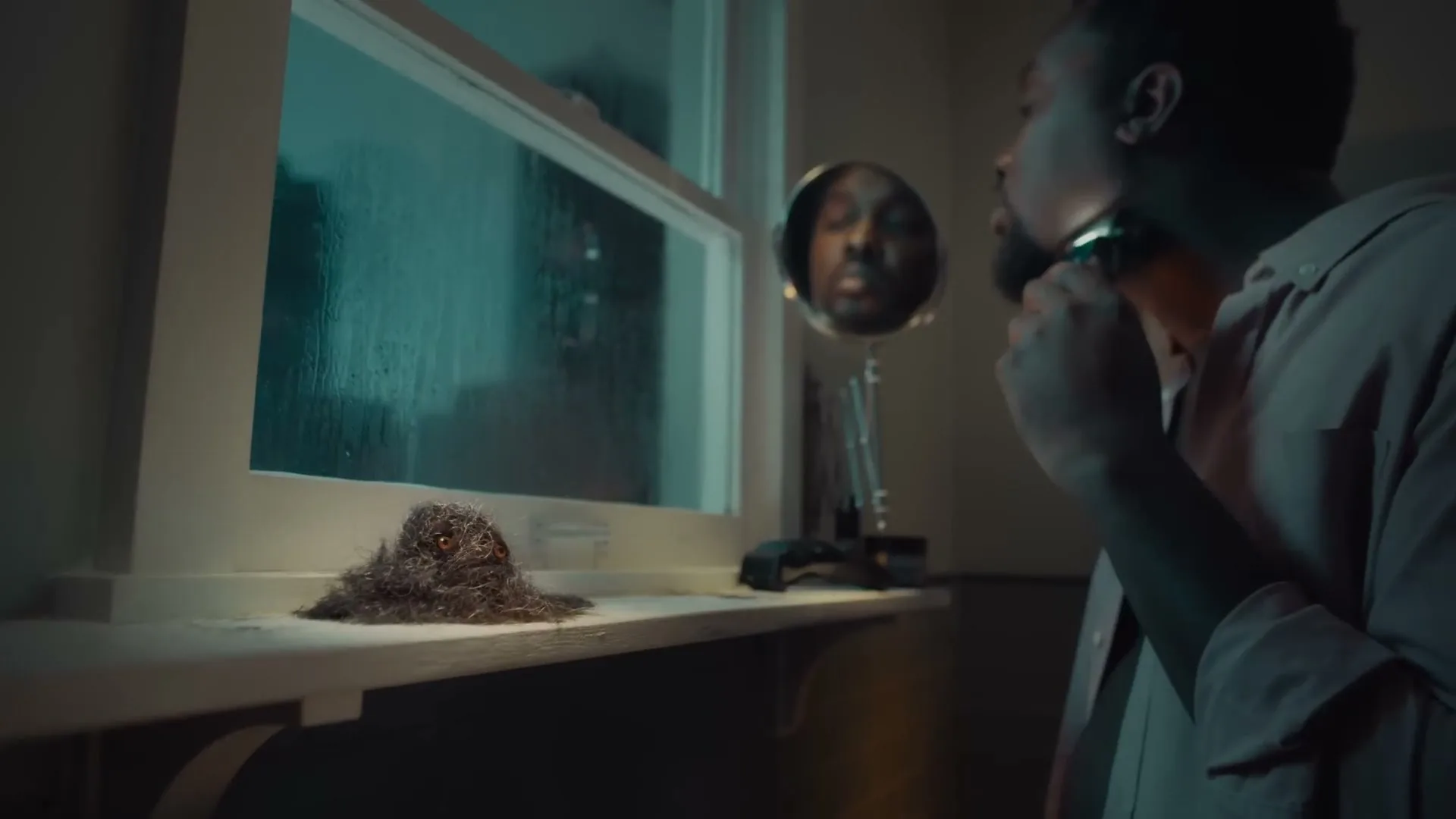 A man shaves his face in a dimly lit bathroom, while a small, furry creature made of hair sits on the windowsill, looking sad.