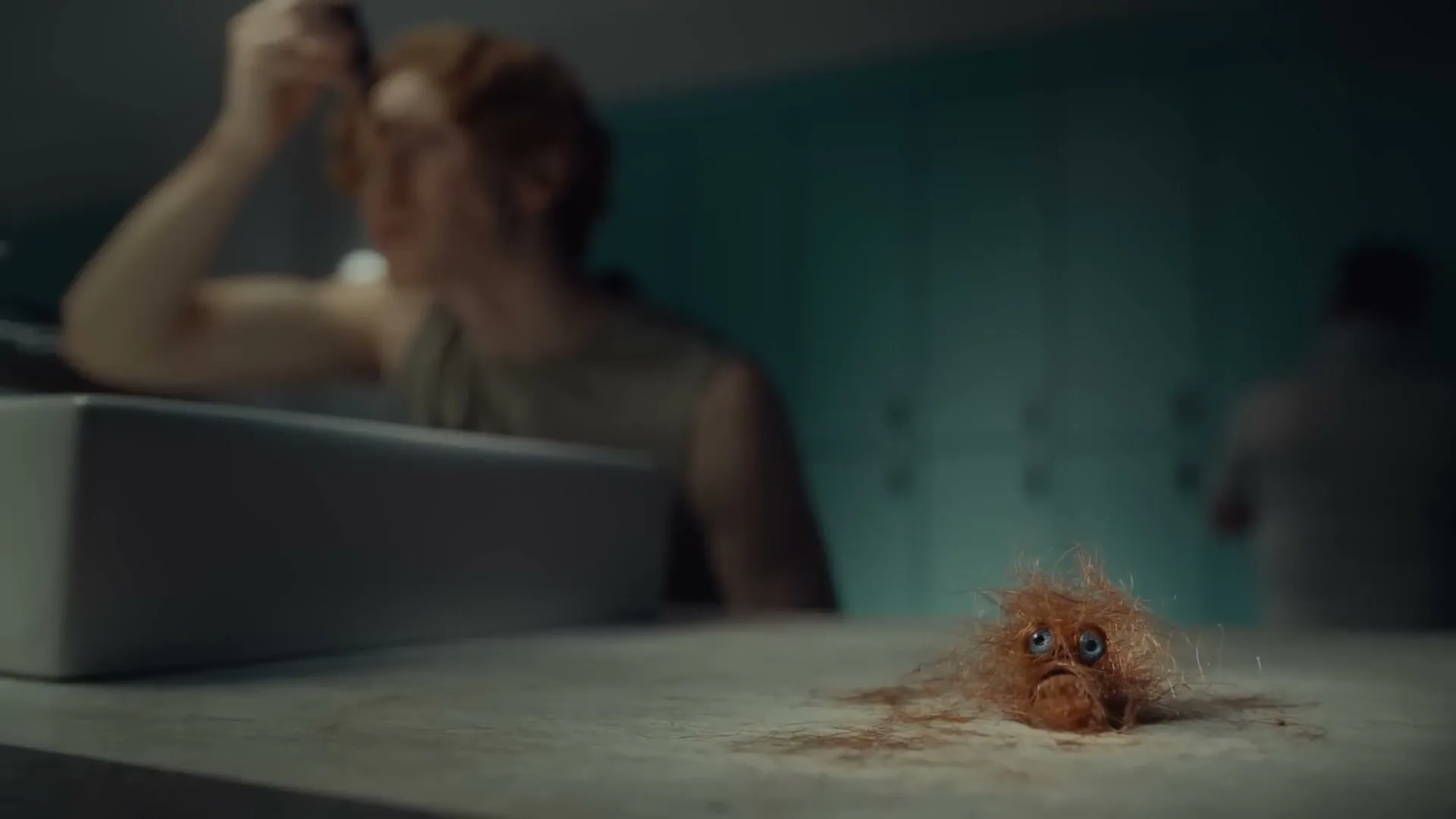 A man with red hair shaves his face in a locker room, while a small, sad, animated hairball sits on the counter.