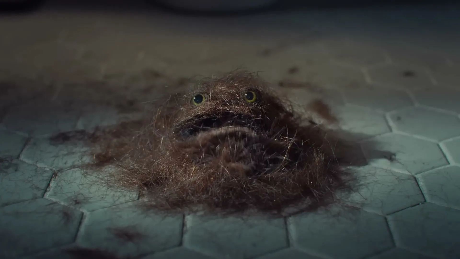 A pile of hair with googly eyes and a mouth, resembling a creature, is on a tiled bathroom floor before being wiped away by a hand with a tissue.