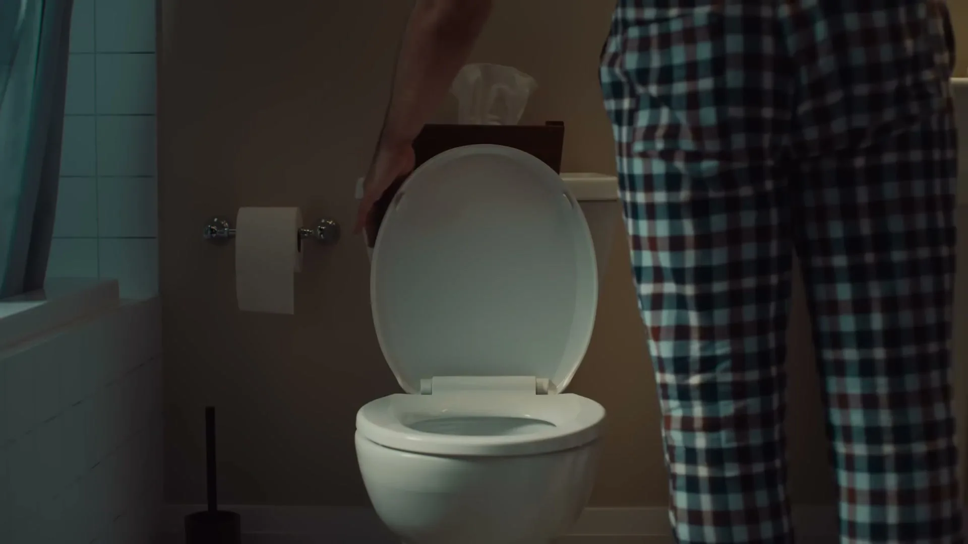 A man in plaid pajama pants drops a tissue into a toilet in a dimly lit bathroom.