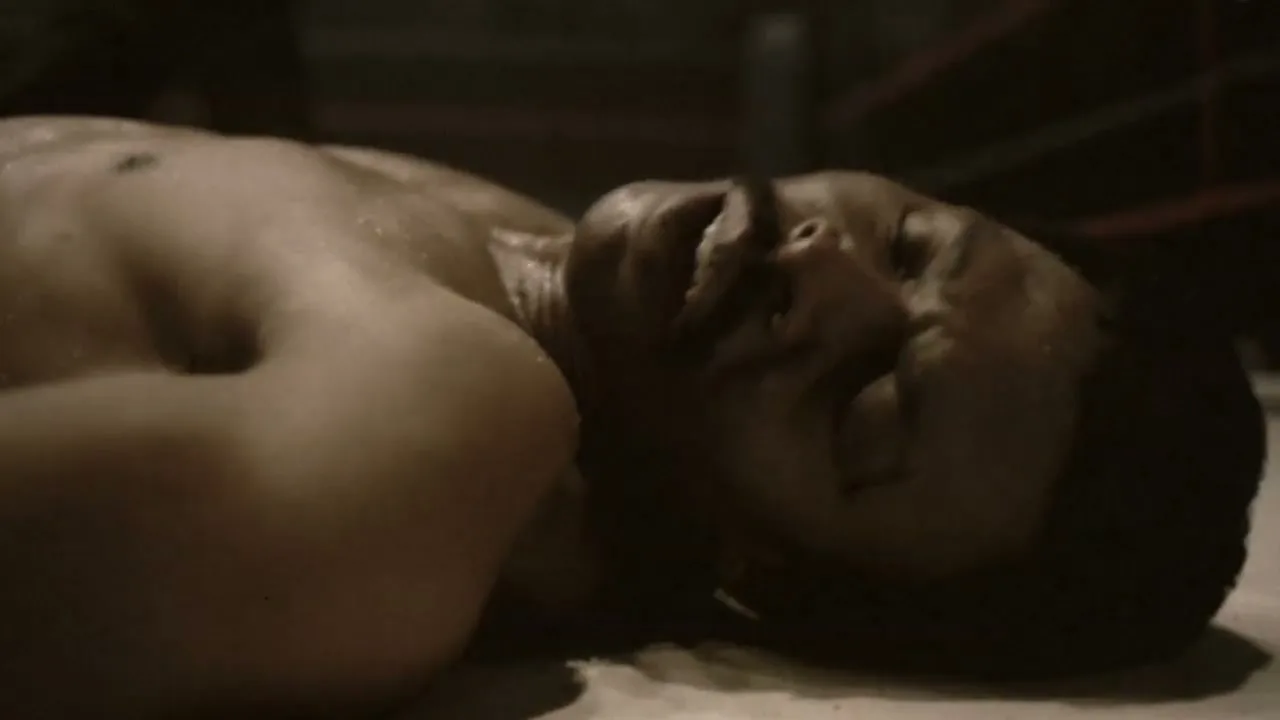 A male boxer, sweaty and shirtless, lies unconscious on the canvas of a boxing ring after being knocked out.