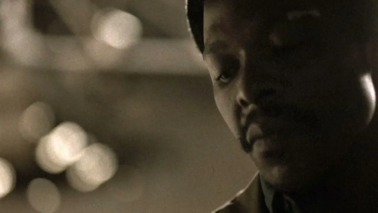 A man with dark skin and a mustache looks down, his face partially obscured by shadow, in a dimly lit interior.
