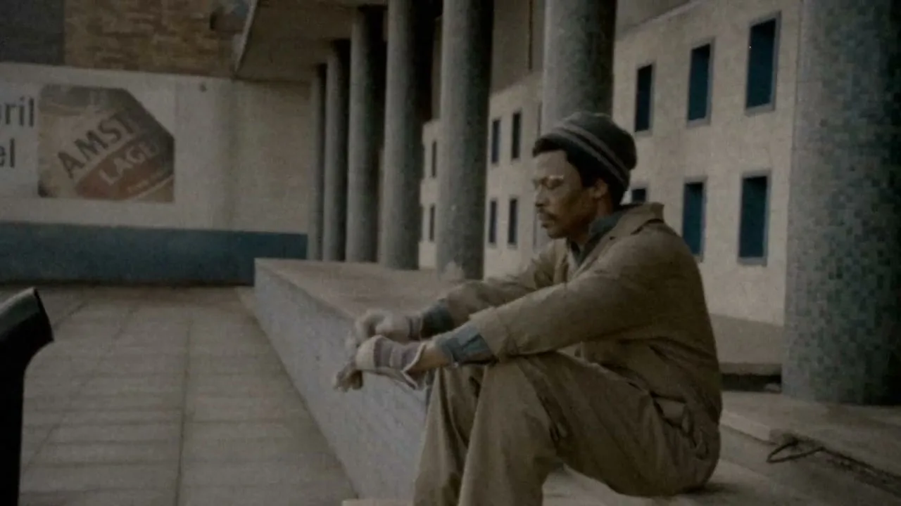 A man in his 40s, wearing a beanie and work clothes, sits alone on a concrete ledge, looking downcast in an empty, industrial-looking space.