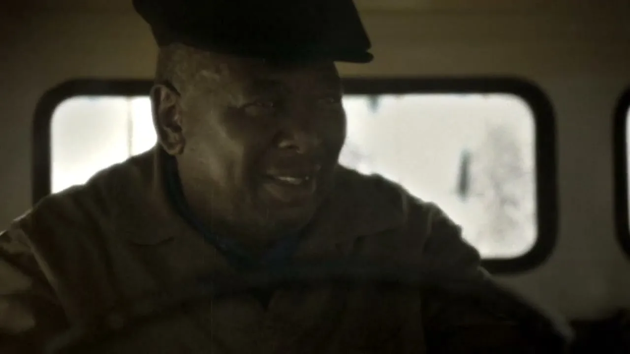 A man in his 60s, wearing a cap and work clothes, smiles while driving a vehicle, with bright windows in the background.