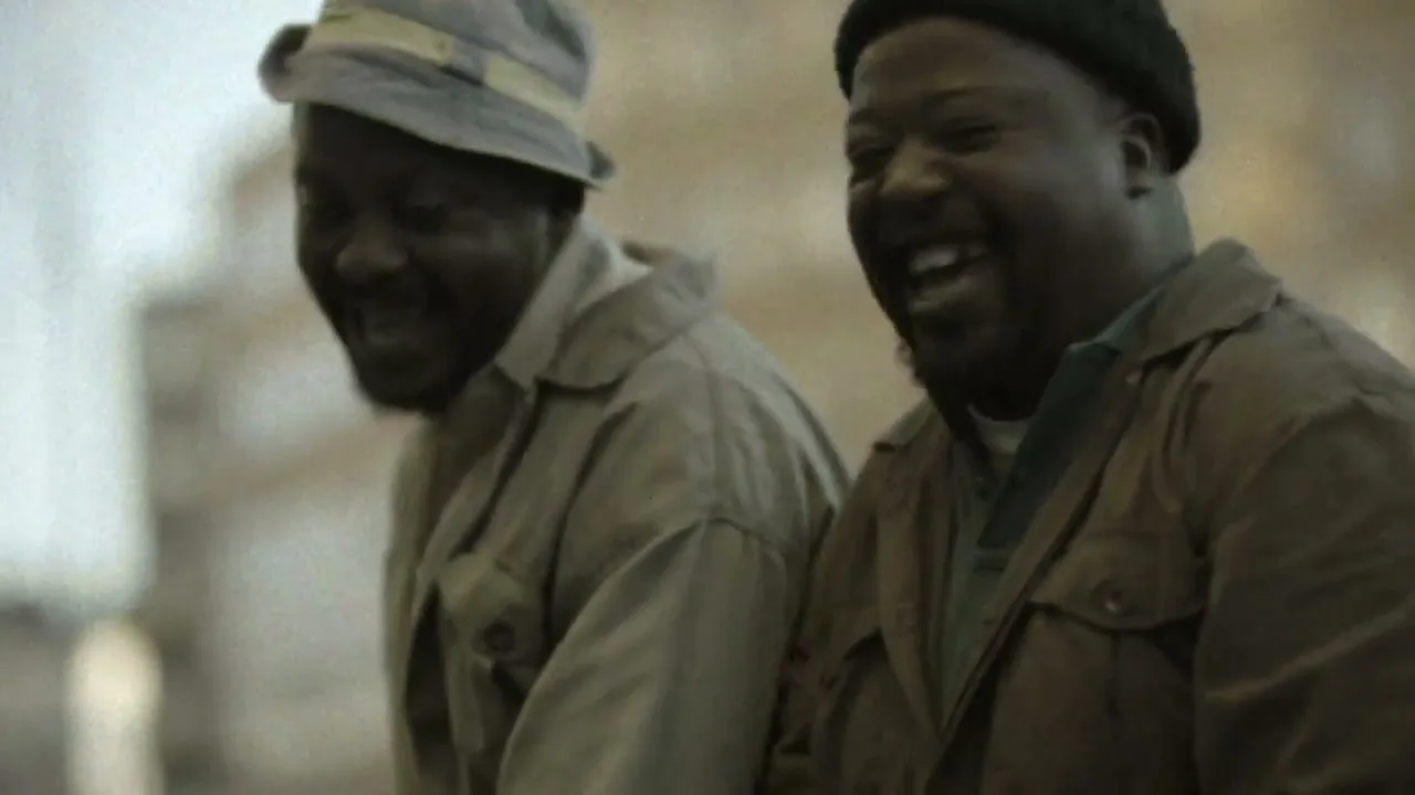 Two middle-aged Black men, one in a bucket hat and the other in a beanie, laugh heartily while looking off-camera in an exterior setting.