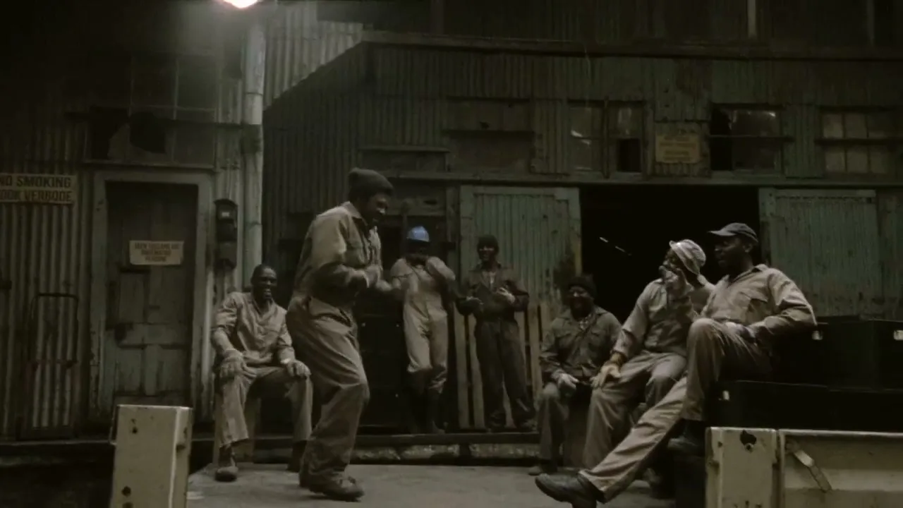 A group of men in work clothes watch one man shadow box in front of a corrugated metal building.