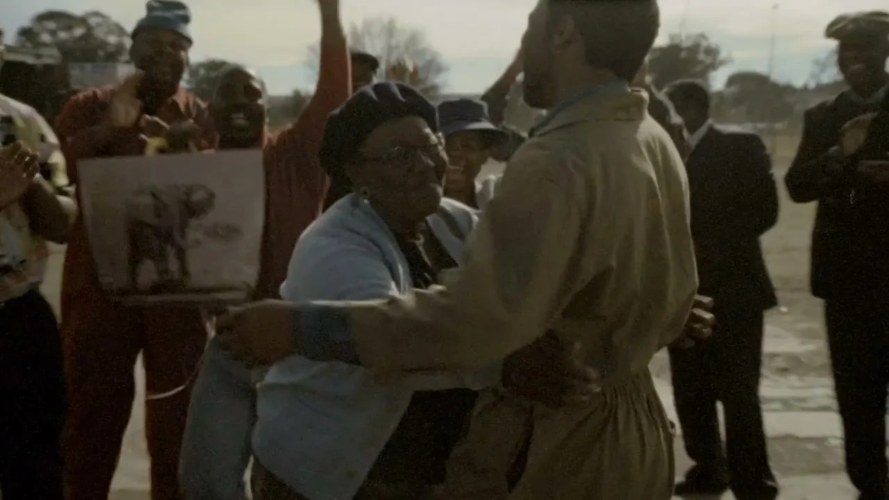 An older woman in a light blue cardigan embraces a man in a tan jumpsuit, surrounded by a cheering crowd outdoors.