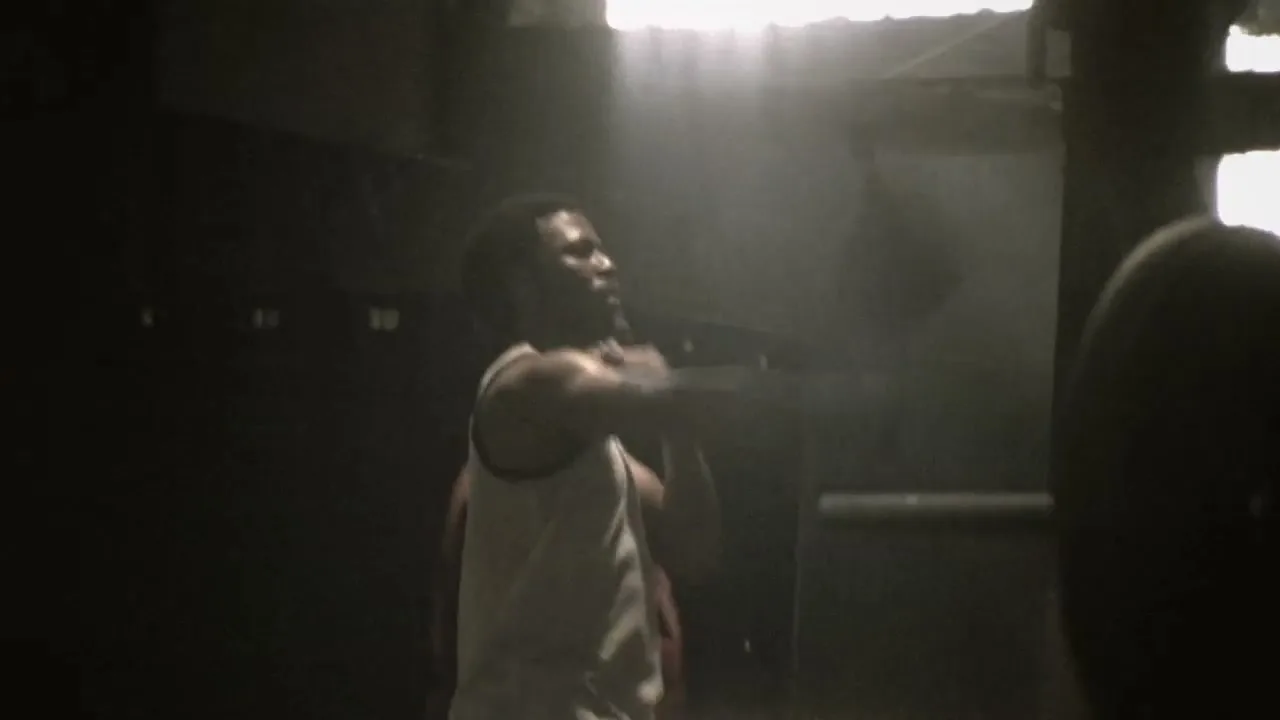 A male boxer in a white tank top trains in a dark, smoky gym, throwing punches as light streams in from above.