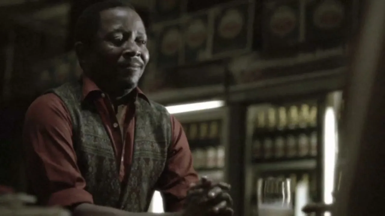 A man in a red shirt and patterned vest sits at a bar, looking up and smiling, with beer bottles in a fridge behind him.
