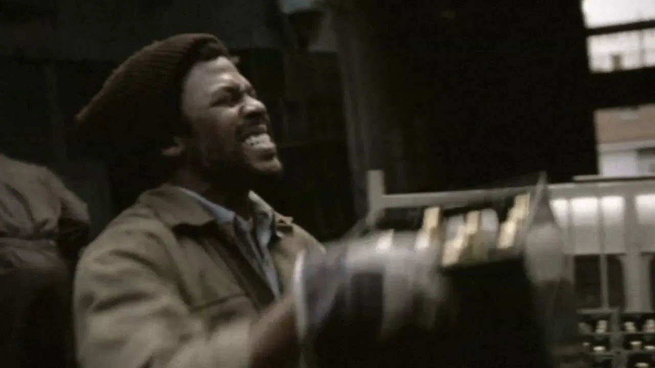 A man in a brown jacket and beanie struggles to lift a heavy object, grimacing with effort in a dimly lit industrial space.