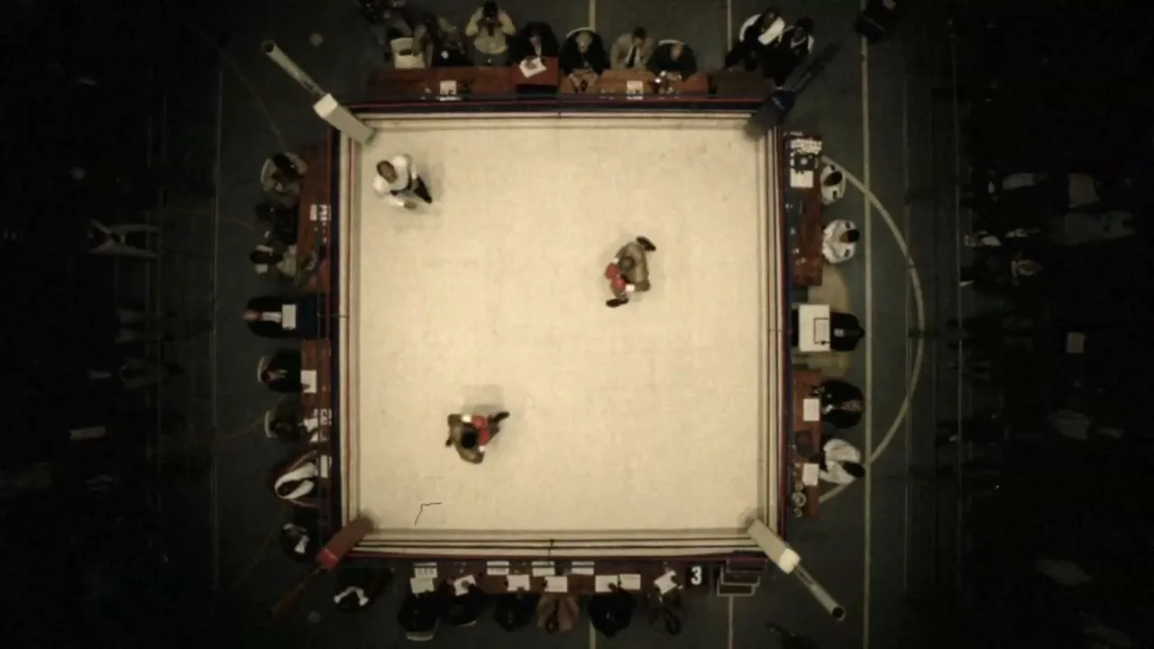 An overhead shot shows two boxers and a referee in a boxing ring, surrounded by spectators in a dimly lit arena.