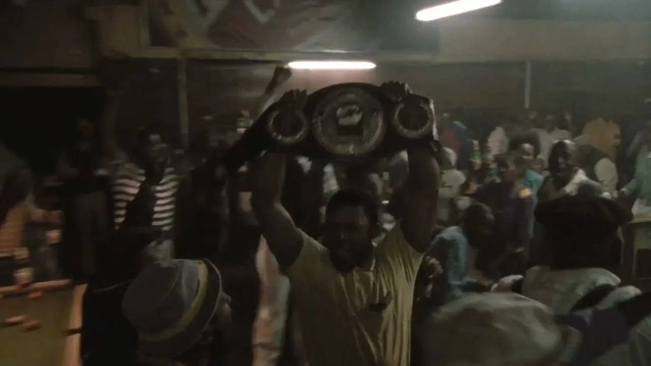A male boxer in a yellow shirt holds up a championship belt in a crowded, dimly lit room, celebrating his victory.