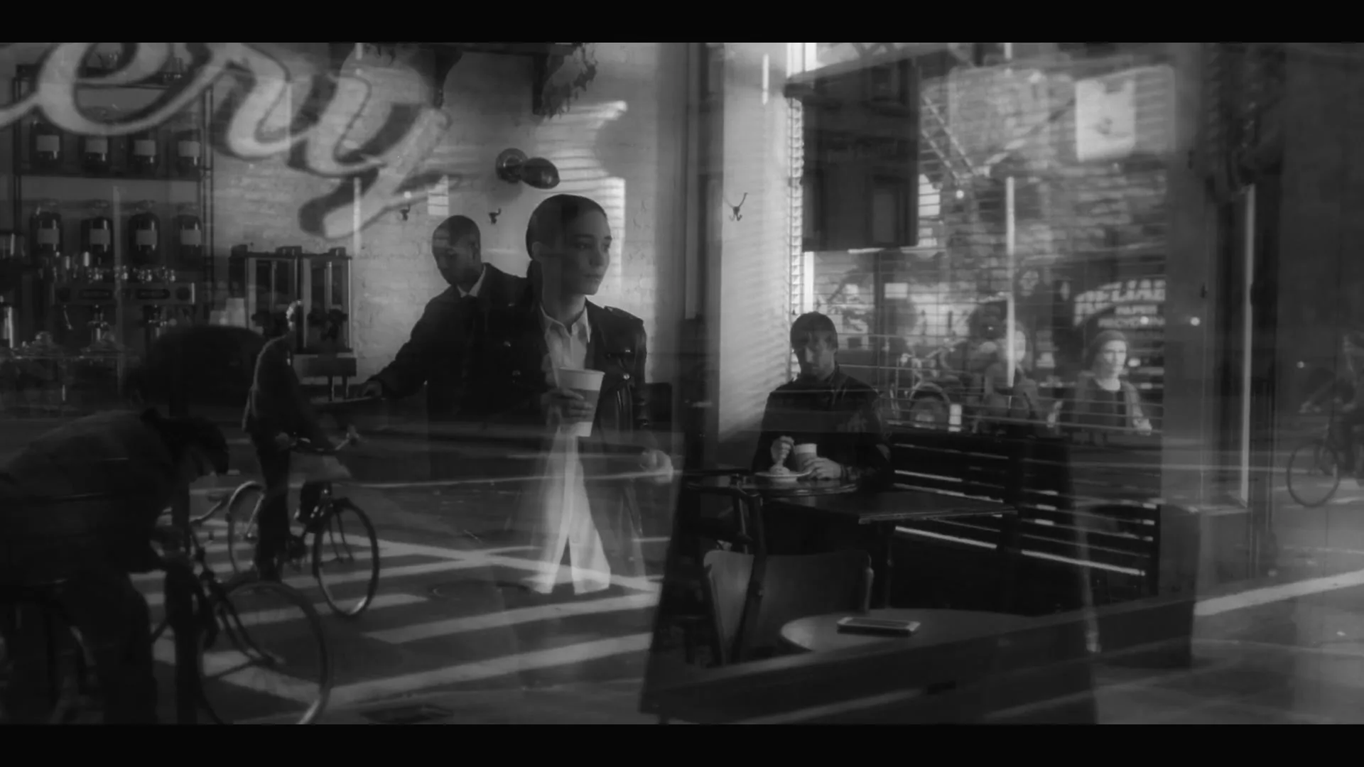 A woman in a white shirt and dark jacket stands in a cafe, holding a cup, with reflections of the street and other people visible in the window.