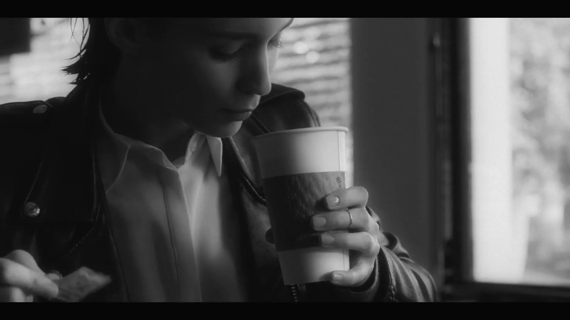 A young woman with short hair, wearing a leather jacket, holds a coffee cup and looks down, possibly eating, in an interior setting.