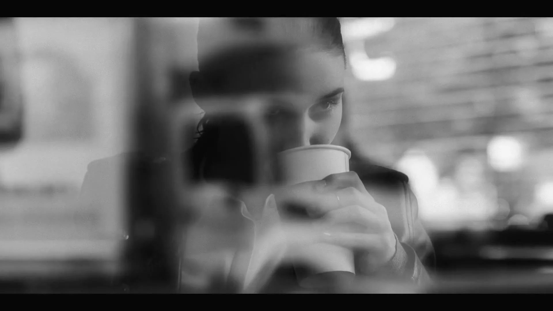A young woman in a leather jacket sips from a coffee cup, looking out a window with reflections, in a black and white shot.