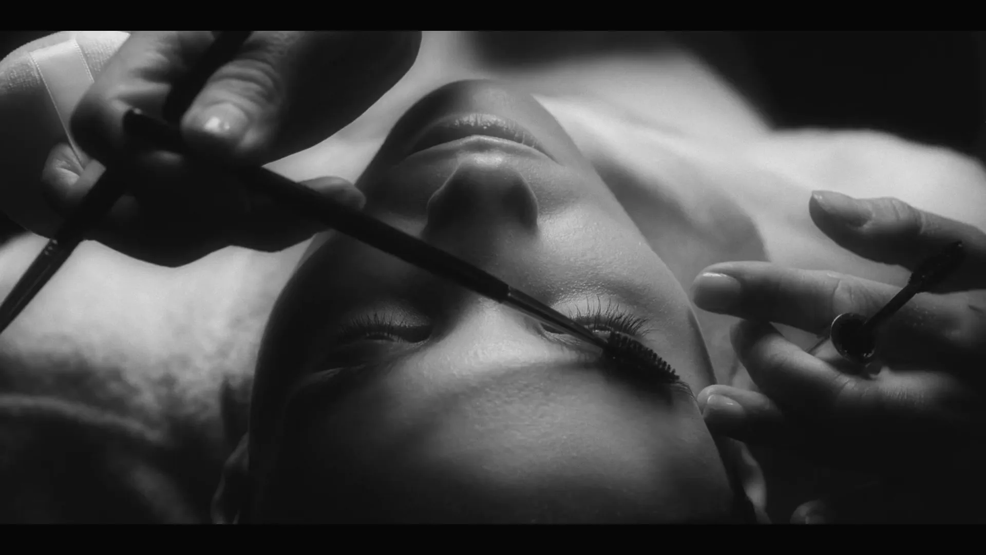 A woman lies down as a makeup artist applies mascara to her eyelashes, in a black and white, high-contrast shot.
