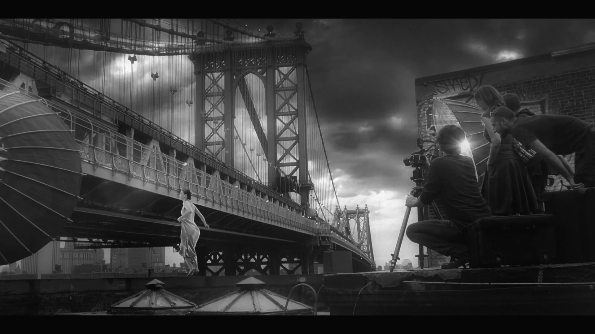 A black and white shot shows a woman in a dress posing on a rooftop with the Manhattan Bridge in the background, while a film crew works nearby.