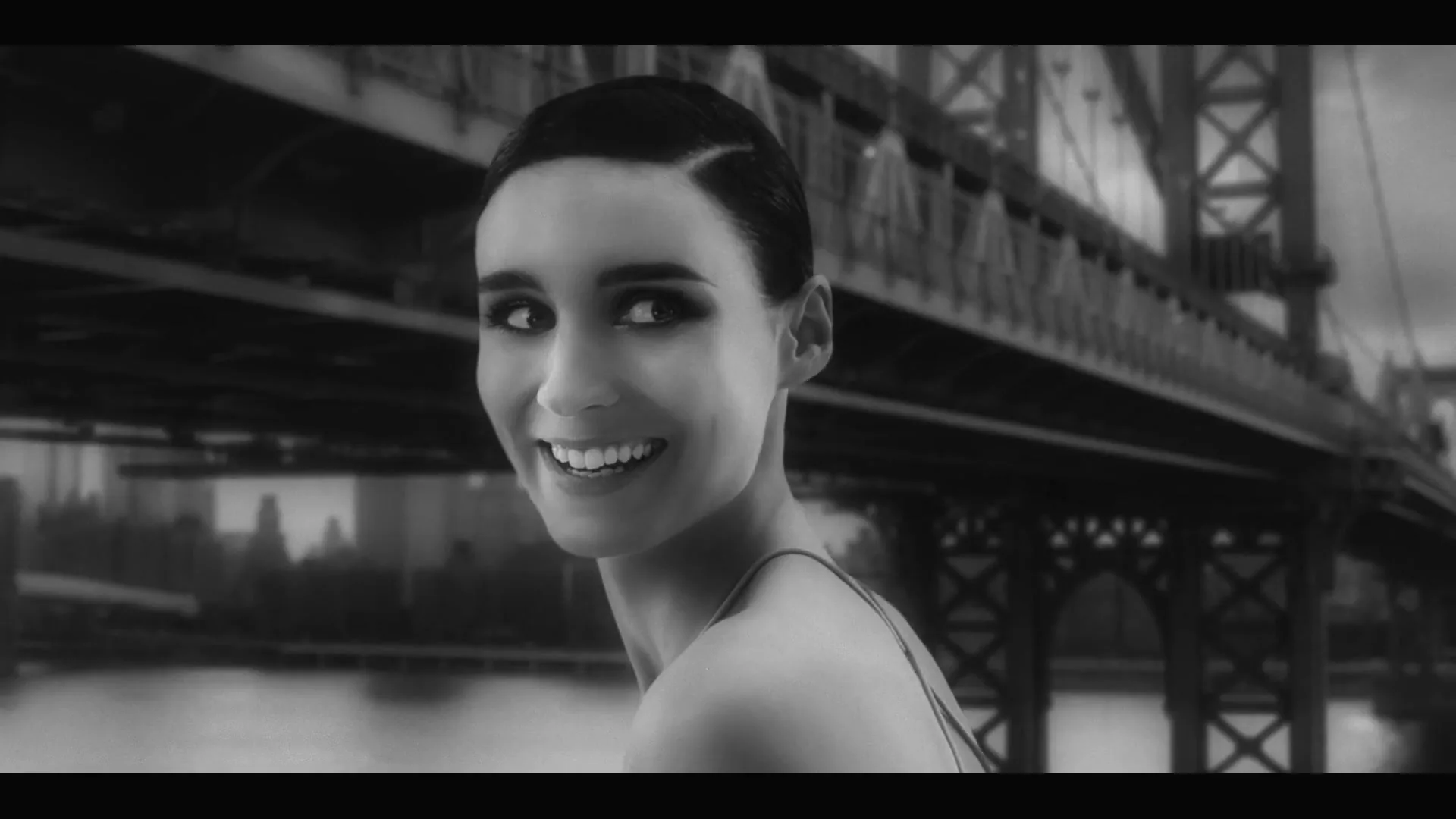 Rooney Mara, a young woman with dark hair and a tank top, smiles at the camera with a bridge in the background.