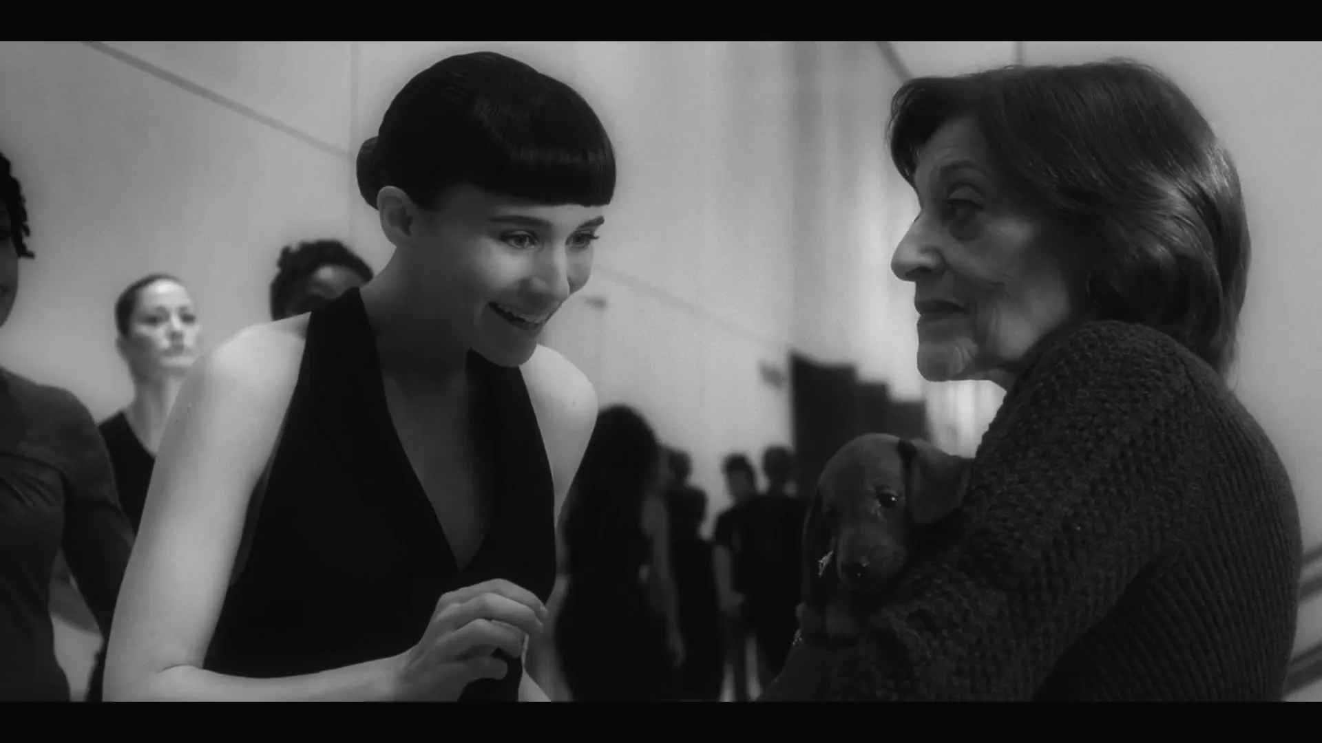 A young woman in a black dress smiles at an older woman holding a small dog in a bright, mirrored room.
