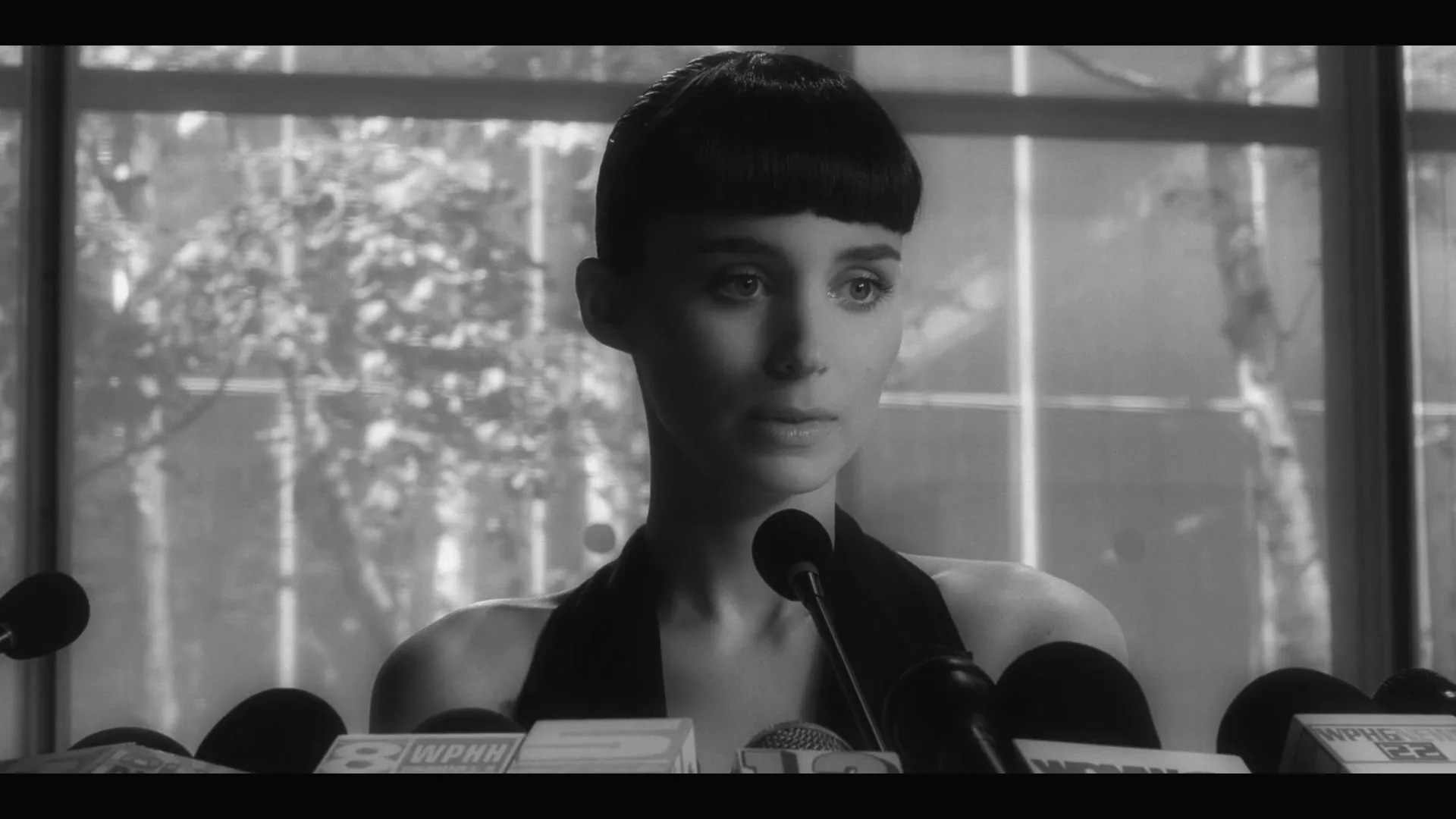 A young woman with short dark hair and a black dress sits at a press conference, looking slightly to the right, surrounded by microphones.