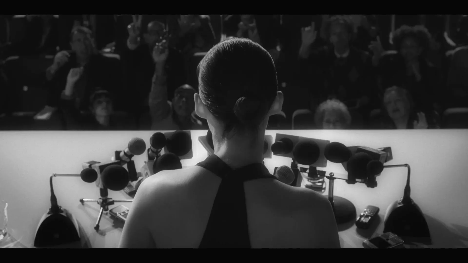 A woman with her hair in a bun, wearing a halter top, sits at a table with many microphones, facing a crowd of people with raised hands.