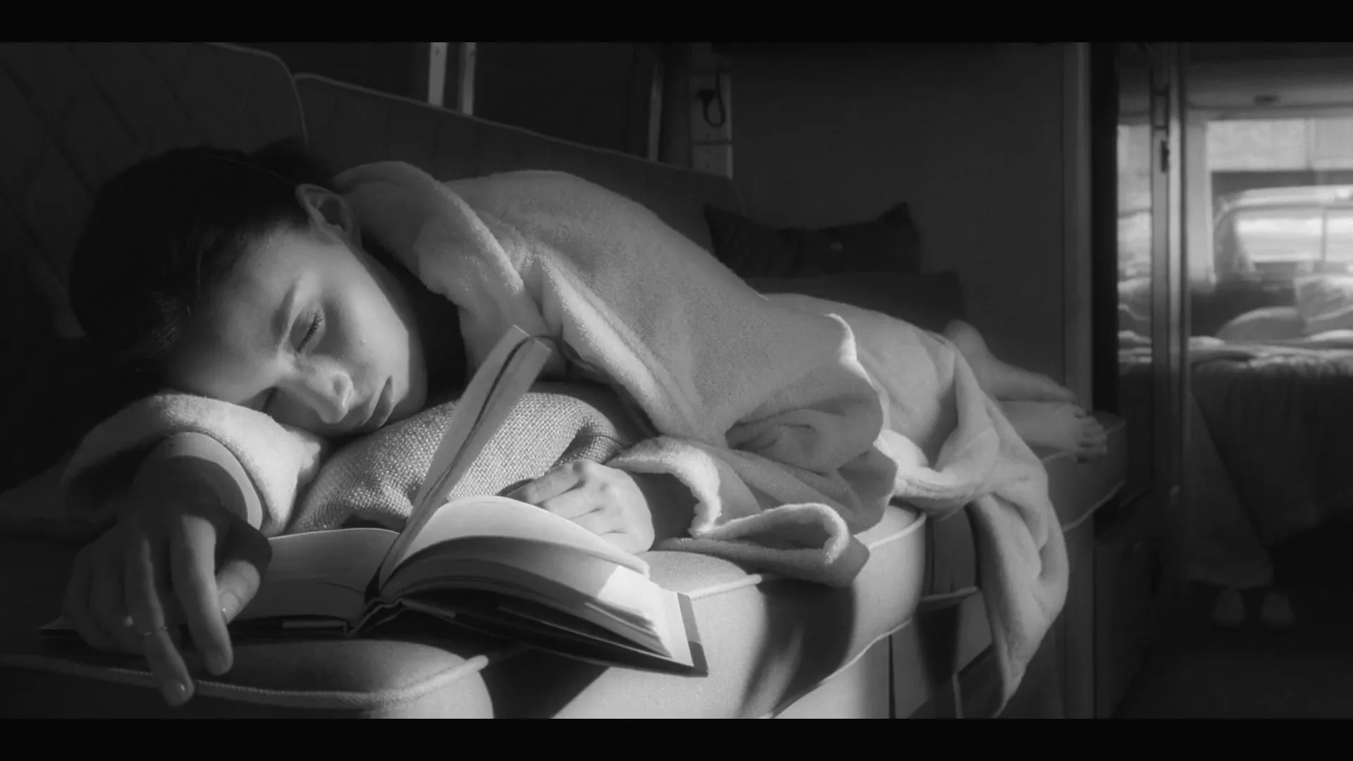 A young woman in a bathrobe is sleeping on a couch with an open book on her chest, bathed in sunlight in a cozy interior.