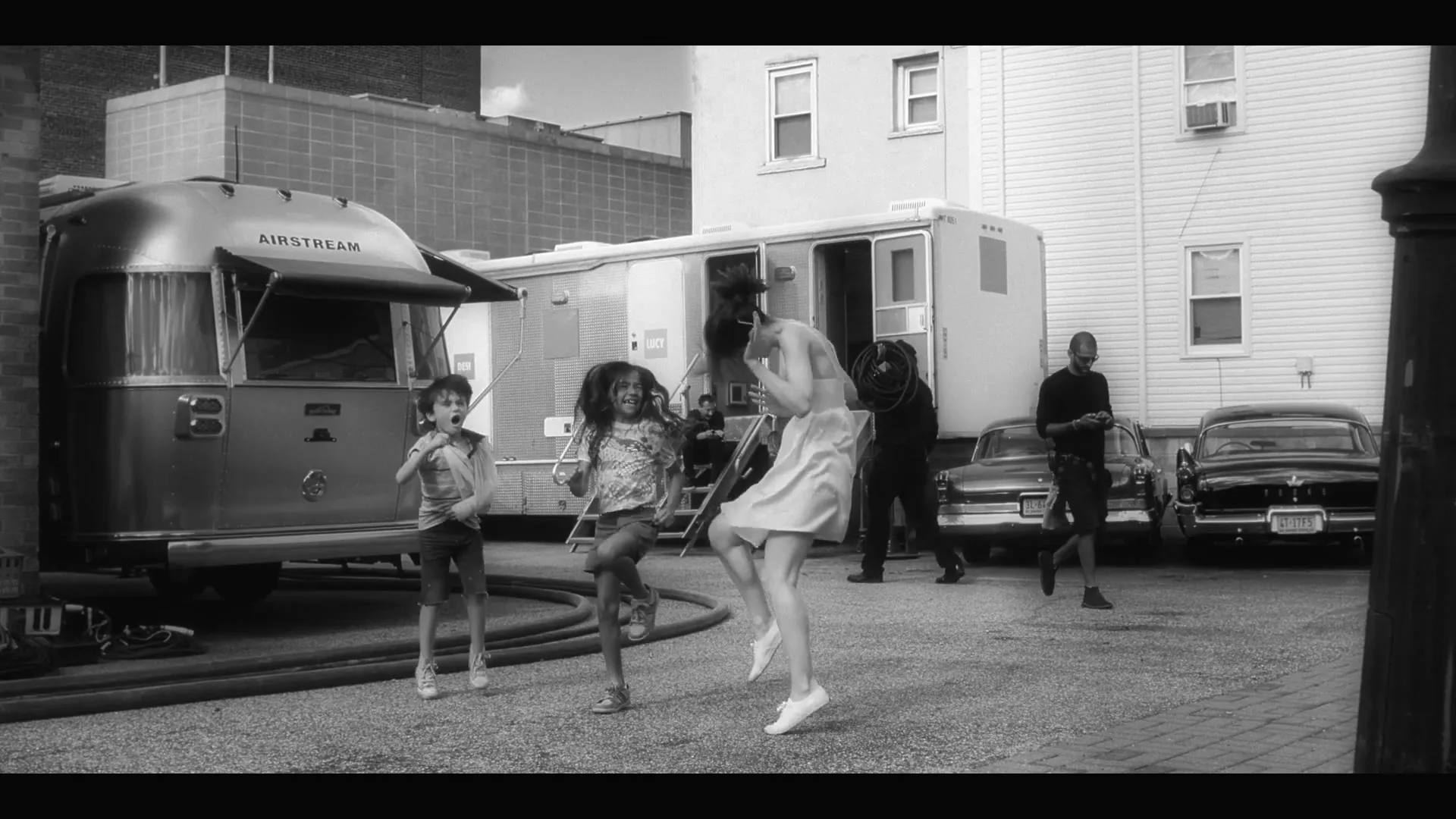A woman in a dress dances with two children in a paved alleyway, with trailers and classic cars in the background.
