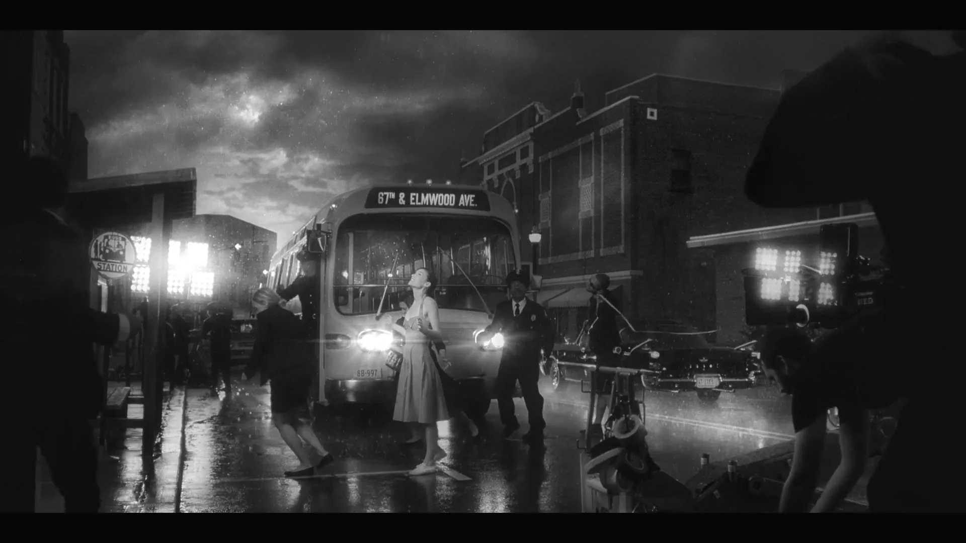A woman in a dress stands in the rain on a city street at night, surrounded by people and a bus, in a black and white shot.