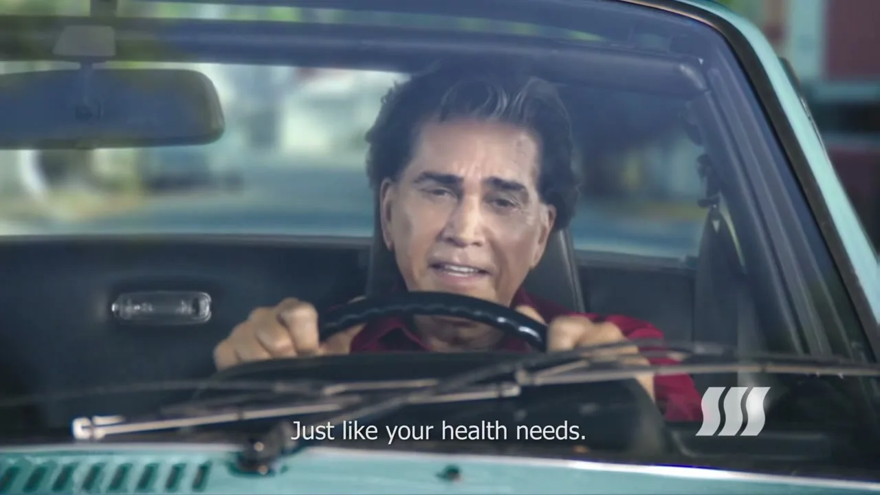 A man with dark hair and a red shirt drives a vintage convertible car, looking forward with a slight smile as text appears on screen.