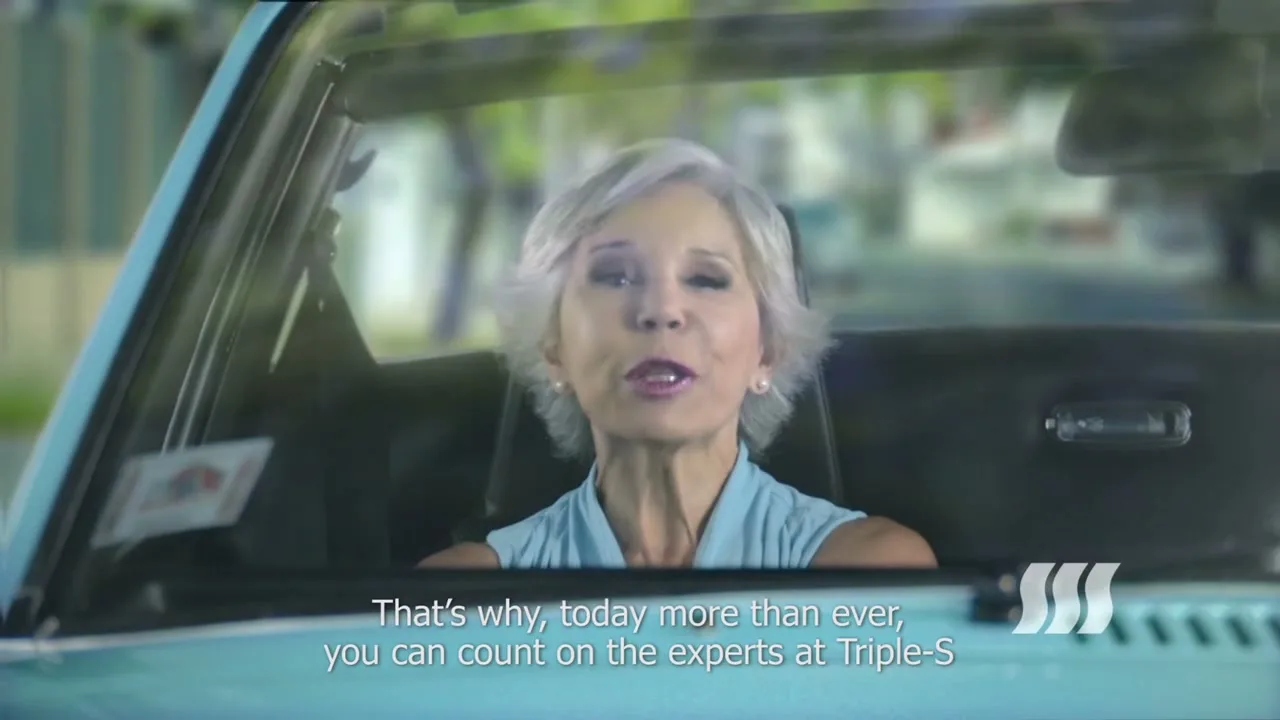 A senior woman with short grey hair, wearing a light blue top, is driving a vintage convertible car and talking to the camera on a sunny day.