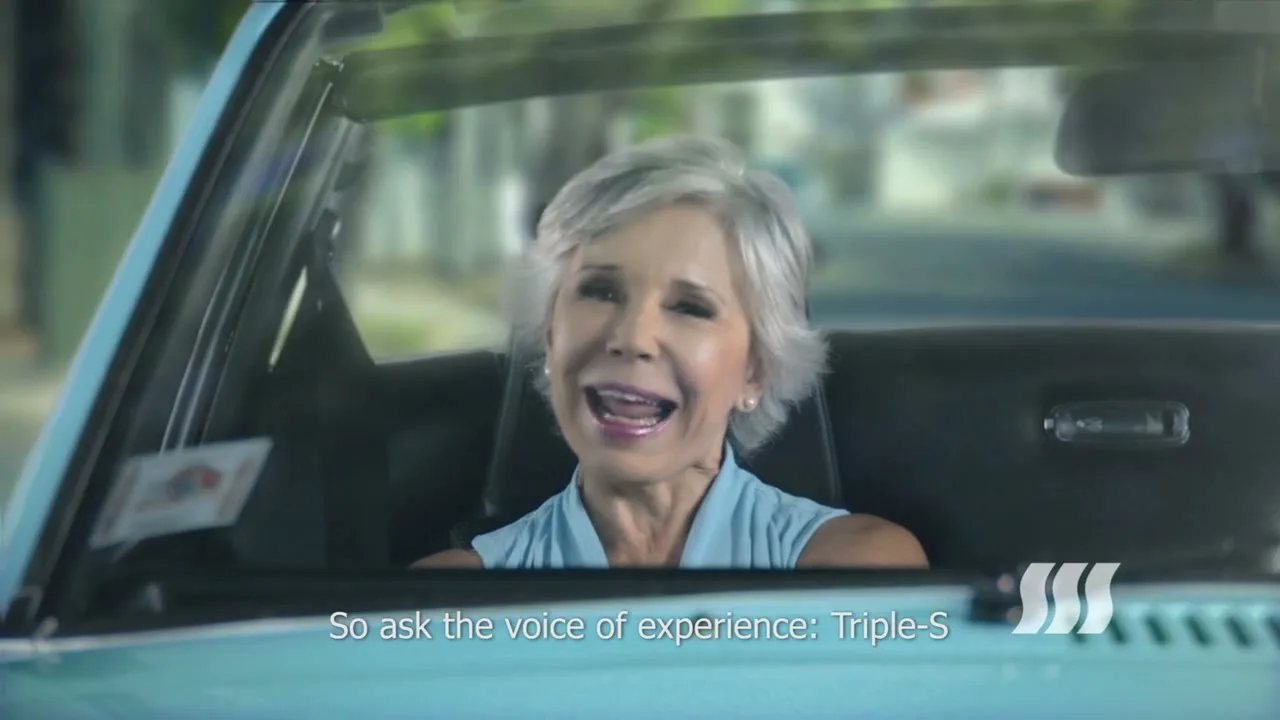 A senior woman with short grey hair, wearing a light blue top, smiles and talks while sitting in the driver's seat of a vintage light blue convertible car on a sunny day.