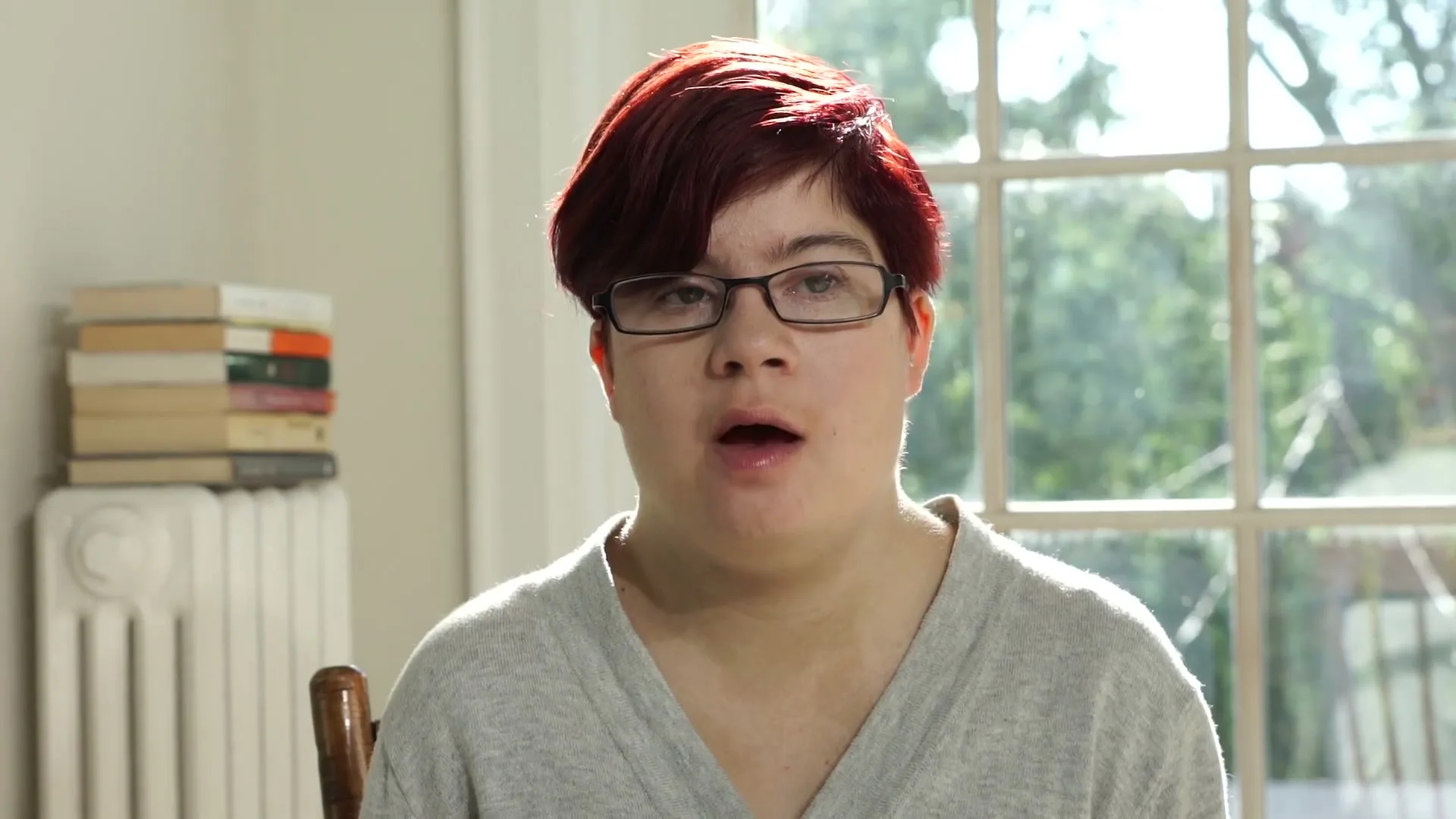 A young woman with short red hair and glasses looks directly at the camera, speaking in a brightly lit room with a window in the background.