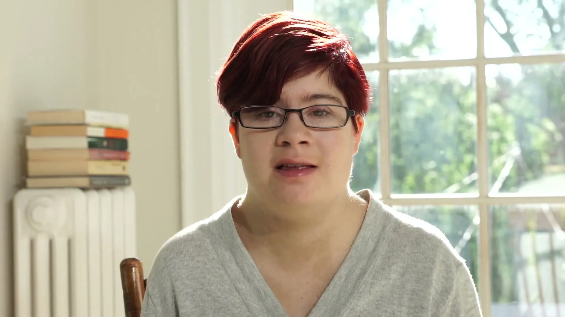 A young woman with short red hair and glasses speaks directly to the camera in a brightly lit room with a window in the background.